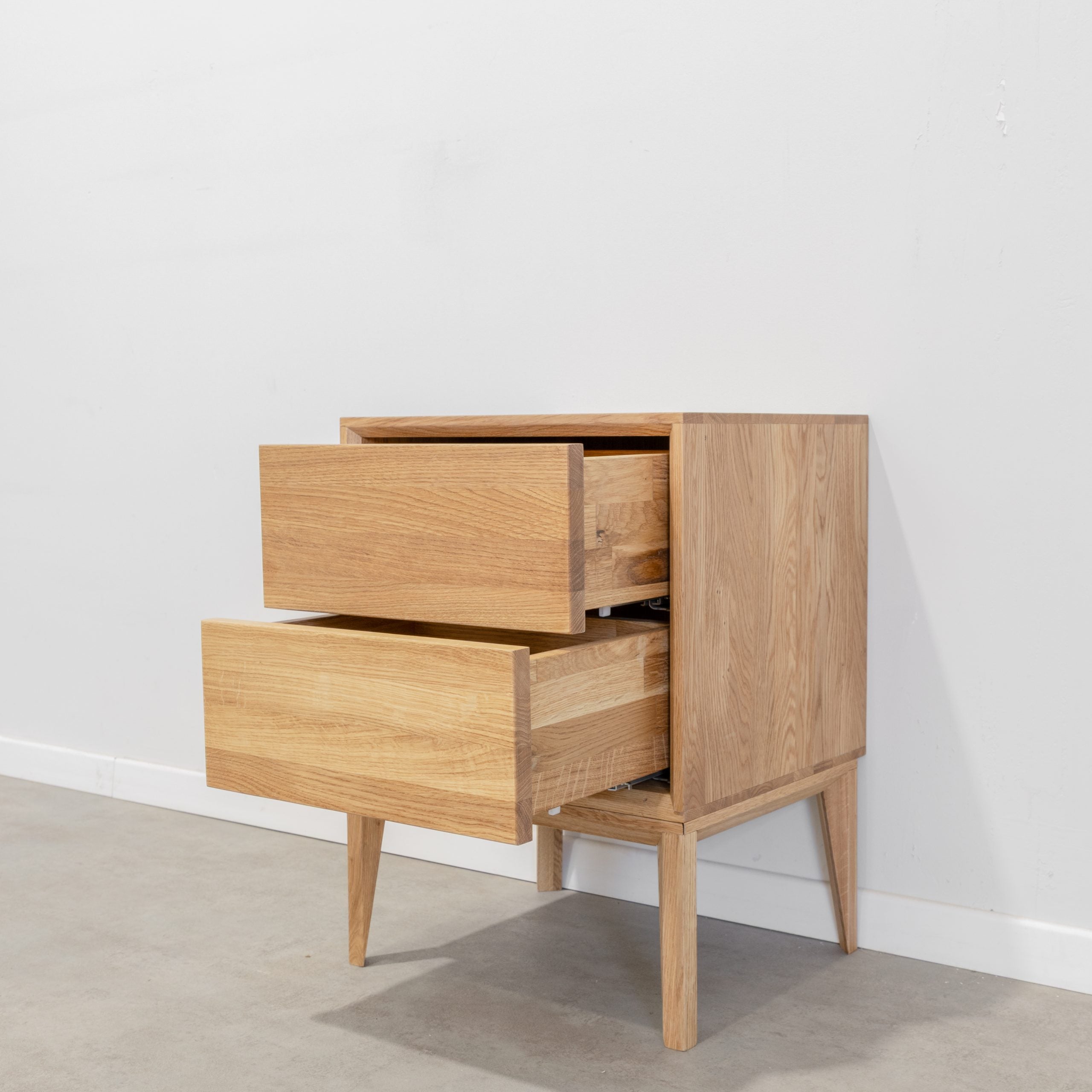 שידת לילה מדגם קודומה עם שני מגירות פתוחות, עומדת על ארבע רגליים ליד קיר לבן ורצפה אפורה.